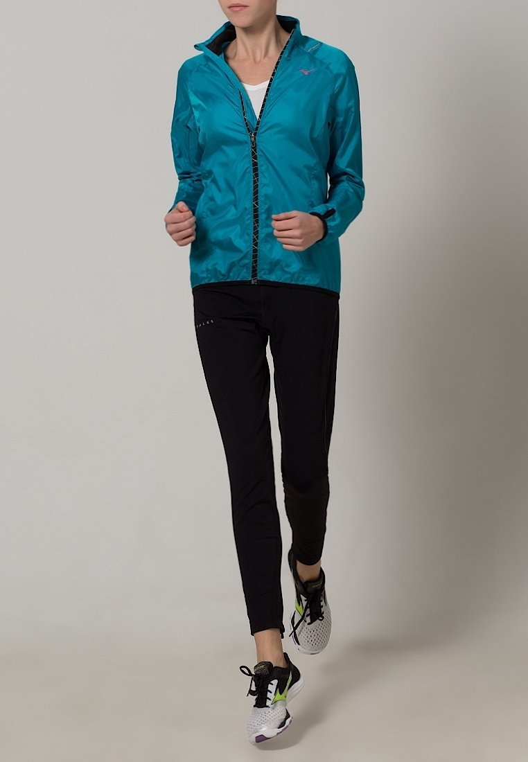 Femme faisant du jogging portant une veste turquoise zippée, des leggings de sport noirs et des chaussures de course blanches avec des accents noirs et vert fluo.