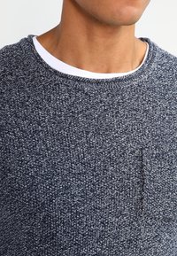 Granatowy sweter z dzianiny z teksturowanym wzorem i małą kieszonką na piersi. Dekolt ma widoczną białą podszewkę.