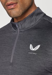 Harmaa vetoketjullinen urheilupaita, jossa on teksturoidu pinta, pieni valkoinen logo rinnassa ja korkea kaulus.