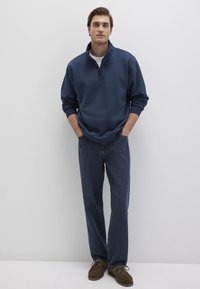 Dunkelblaues Zip-Pulli mit langen Ärmeln, kombiniert mit geraden dunklen Jeans und braunen Wildlederschuhen. Einfaches, lässiges Design.