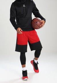 Persona con sudadera negra con capucha, pantalones cortos de baloncesto rojos y negros, y zapatillas rojas sosteniendo un balón de baloncesto con el logo de Jumpman.
