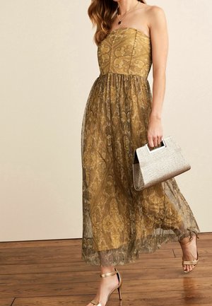 Mujer con vestido sin tirantes de patrón dorado, sosteniendo un bolso beige con textura y caminando con sandalias de tacón doradas sobre un suelo de madera.