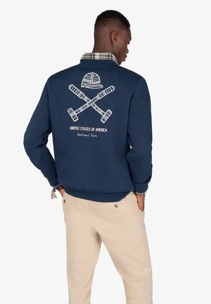 Hombre vistiendo un sudadera azul marino con un gráfico de palas de canoa cruzadas y un sombrero, y el texto "Parque Nacional de los Estados Unidos de América" en la parte posterior.