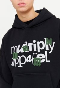 Sudadera negra con el texto blanco "multiply apparel" y tres letras bordadas en verde. Cuenta con un bolsillo canguro y capucha con cordón.