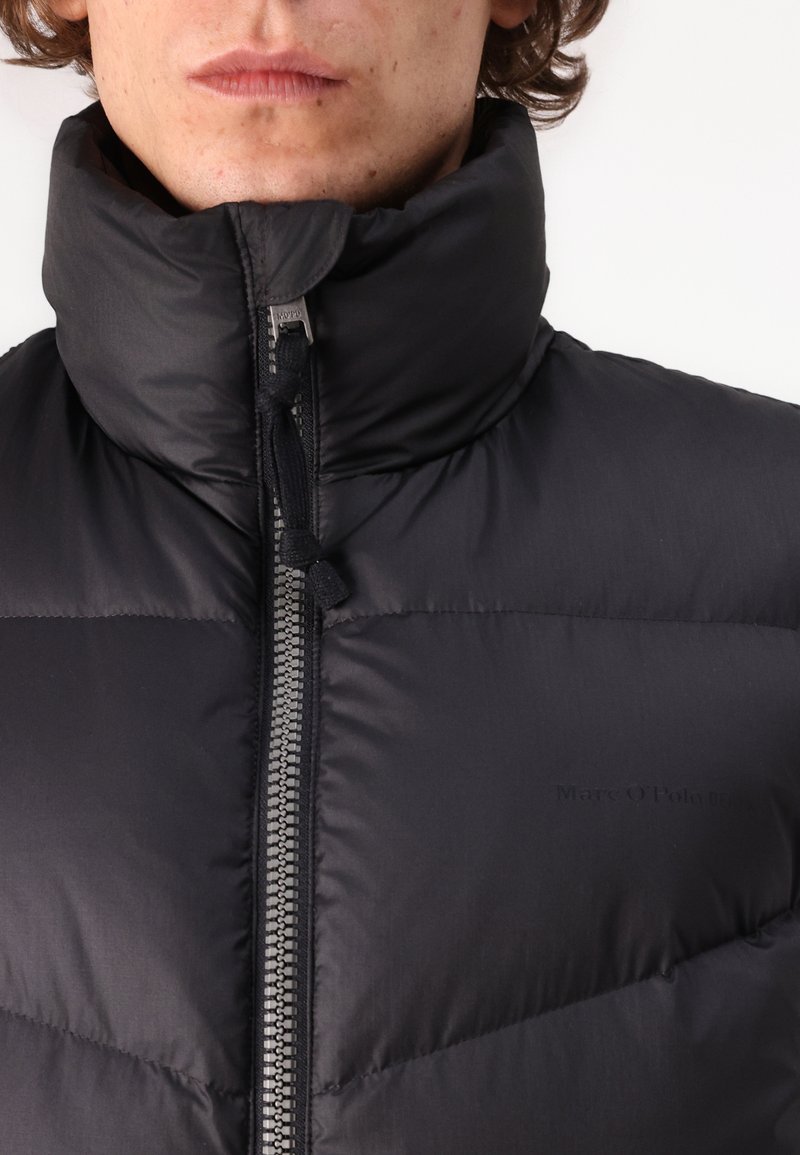 Veste rembourrée noire avec un col montant, dotée d'une fermeture éclair à l'avant et d'une languette de tirage. Le tissu a une texture lisse, avec un logo imprimé sur la poitrine.