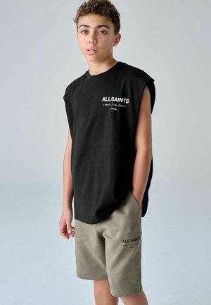 Noor lühikeste lokkis juustega, kandes musta varrukateta AllSaintsi särki ja oliivirohelisi AllSaintsi lühikeseid pükse, seismas ühe käega pihus.