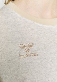 Lichtgrijze T-shirt met een beige geribbelde kraag; heeft een reliëflogo met een bijenontwerp en de merknaam "hummel."