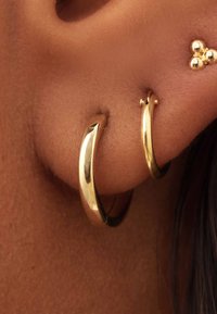 Des boucles d'oreilles créoles en or exposées sur la peau, avec une surface lisse et polie. Une créole est plus grande, légèrement courbée, avec une plus petite créole au-dessus.