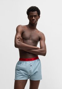 Lyseblå shorts med en rød "HUGO" talje, der har knaplukning, glat tekstur og en afslappet pasform. Tilbehøret inkluderer en sølv halskæde.