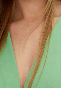 Prenda verde con un profundo escote en V. Textura suave, tela ligera. Mechas de cabello rubio parcialmente visibles en el lado izquierdo.