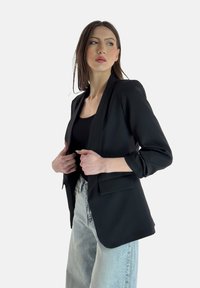 Blazer nero realizzato in un tessuto liscio con chiusura a singolo bottone, rever a notched e due tasche frontali. Indossato sopra un top nero.