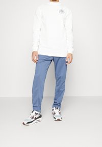 White long-sleeve shirt with a logo, paired with blue pants and multicolored sneakers featuring black, white, and burgundy accents.