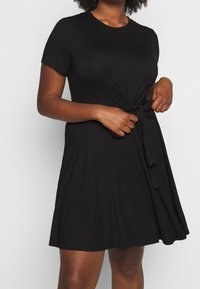 Robe noire à manches courtes avec un col rond et un détail de nouage à l'avant au niveau de la taille, fabriquée à partir d'un tissu lisse et extensible. Silhouette évasée.
