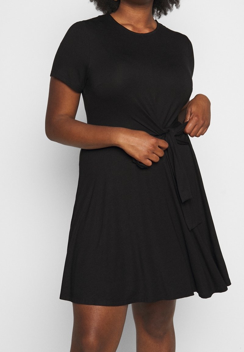 Robe noire à manches courtes avec un col rond et un détail de nouage à l'avant au niveau de la taille, fabriquée à partir d'un tissu lisse et extensible. Silhouette évasée.