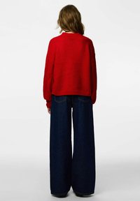 Czerwony sweter na drutach o luźnym kroju z prążkowanym wykończeniem na dole, zestawiony z szerokimi, ciemnymi jeansami o wysokim stanem i tylnych kieszeniach.