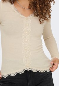 Vaaleanbeige ribattu pitkähihaisuus paita, jossa on aallotettu pitsireunus, koriste-elementit ja teksturoitu kangas. Istuu juuri vyötärön alapuolelle.