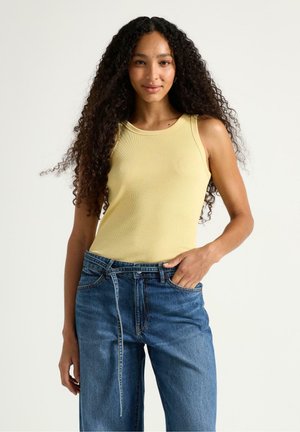 Nainen pitkine kiharine hiuksineen, keltainen hihaton paita ja siniset farkut, seisoo valkoista taustaa vasten, toinen käsi taskussa.