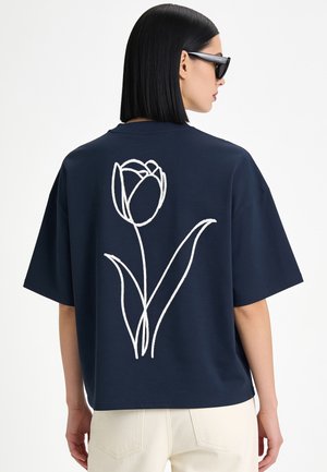 Frau mit schwarzem Bob-Haarschnitt und Sonnenbrille, die ein locker sitzendes dunkelblaues T-Shirt mit einer großen weißen Tulpenlinienzeichnung auf dem Rücken trägt.