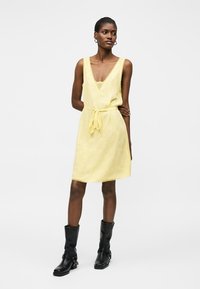 Γυναίκα φορεί αμάνικο κίτρινο φόρεμα με V-λαιμόκοψη και δεμένη μέση, συνδυασμένο με μαύρες μπότες μέχρι τη μέση της γάμπας, στέκεται μπροστά σε λευκό φόντο.