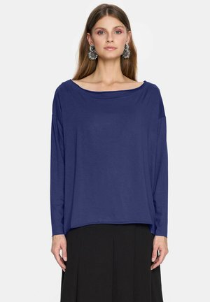 Młoda kobieta w luźnej granatowej bluzce z długim rękawem, czarnej spódnicy oraz dużych srebrnych efektownych kolczykach, stojąca na tle jednolitego tła.