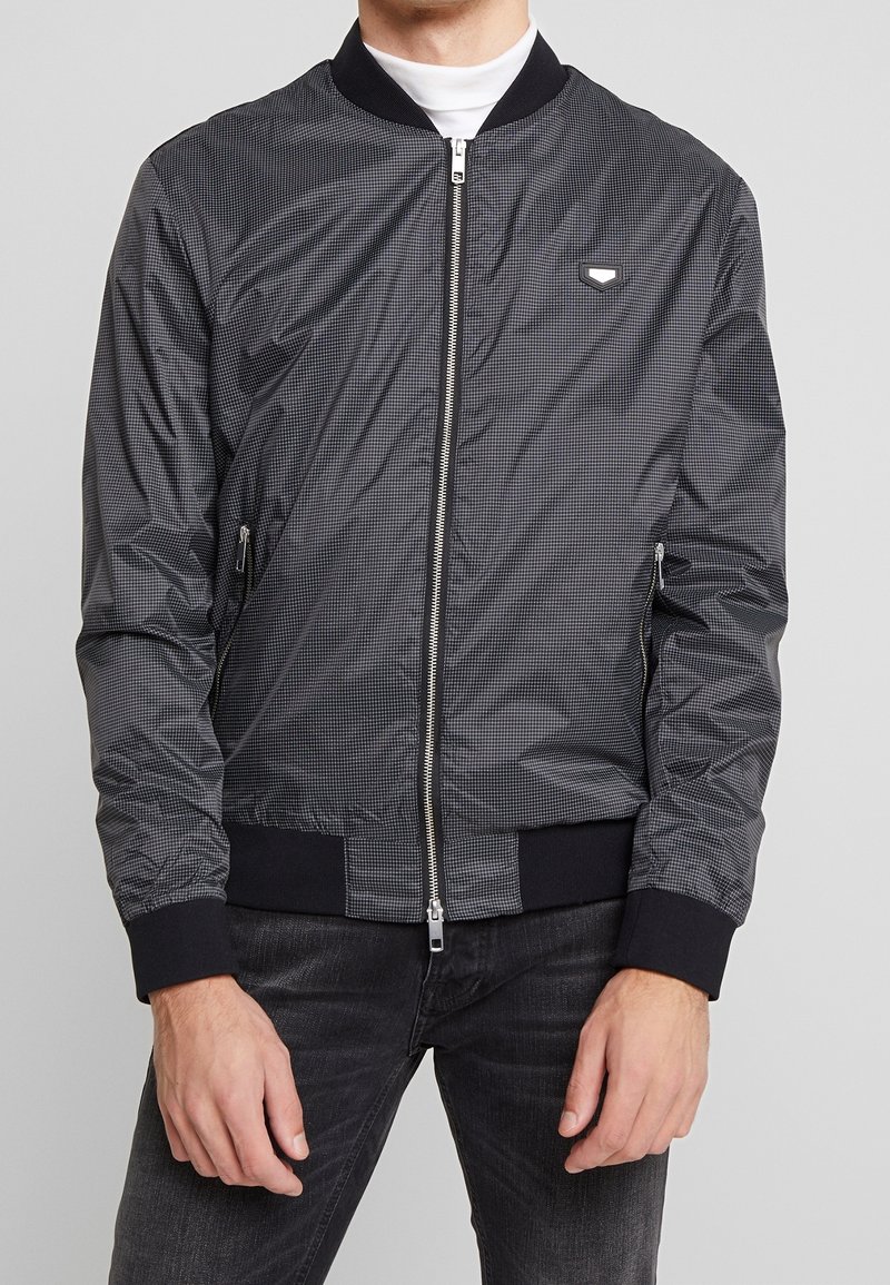 Svart bomberjacka med rutigt mönster, dragkedja, ribbade ärmslut och nederkant, samt sidofickor. Tillverkad av lättvikts, glänsande tyg.