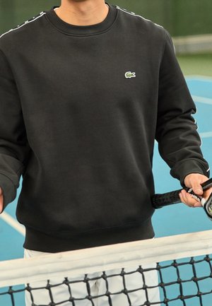 Czarna bluza z okrągłym dekoltem, z małym zielonym logo krokodyla i białymi akcentami na szwach na ramionach, noszona podczas trzymania rakiety tenisowej.