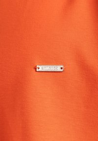 Oranžová tkanina s hladkým povrchem a malou stříbrnou metalickou cedulkou s textem "SMILODOX" připevněnou k ní.