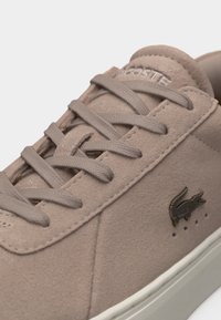 Μπεζ σουέντ αθλητικό παπούτσι με κορδόνια στο ίδιο χρώμα, λευκή σόλα και μεταλλικό λογότυπο κροκόδειλου στο πλάι, που αναδεικνύει τη μάρκα Lacoste στη γλώσσα.