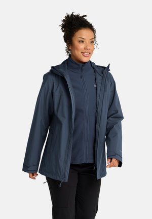 Frau trägt mehrere navyblaue Outdoorjacken übereinander und schwarze Hosen, lächelt und schaut leicht seitlich auf einem einfarbigen hellen Hintergrund.