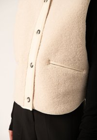 Creme fleece vest med knaplukning fortil, der har sølvknapper og en sidelomme. Den glatte stofoverflade står i kontrast til den bløde fleece.