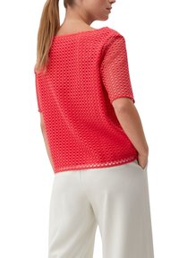 Femme avec une longue queue de cheval, portant un haut texturé corail à manches courtes et un pantalon blanc à jambes larges, les mains dans les poches, vue de dos.