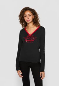 Frau mit lockigem Haar, die ein schwarzes Langarm-V-Ausschnitt-Shirt mit rotem Saum und rotem Text "Replay Custom Specialist Allentown, PA" trägt und steht.