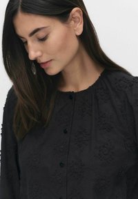 Svart, broderad blus med blommönster, rundad halsringning och knappstängning. Har lång ärm och mjukt tyg.