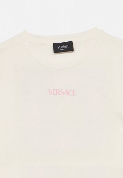 Μπλουζάκι Versace σε κρεμ απόχρωση με ροζ λογότυπο μπροστά και μαύρη ετικέτα με την ένδειξη «Versace Made in Italy» στον λαιμό.