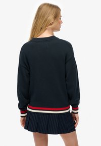 Pull en maille bleu marine avec un ourlet côtelé rayé rouge, blanc et noir. Il présente des manches longues et une coupe décontractée, associé à une jupe plissée.