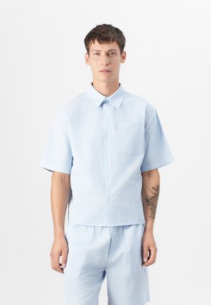 Camisa de manga corta color azul claro con un patrón texturizado, cierre de botones en el frente y bolsillo en el pecho. Combinada con pantalones cortos a juego en azul claro.