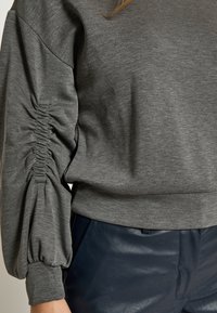 Sudadera gris con detalle fruncido en la manga izquierda, fabricada en tejido suave, con un corte relajado y un dobladillo liso. Combinada con pantalones azul marino.