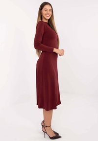 Bordowa sukienka z długim rękawem, z dopasowanym gorsetem i zwiewną spódnicą, wykonana z gładkiego materiału. Noszona z czarnymi szpilkami.
