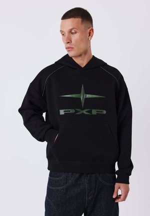 Jeune homme aux cheveux courts portant un sweat à capuche noir avec un logo étoile verte et l'inscription "PXP", les mains dans la poche avant, regardant sur le côté.