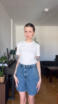 Νεαρή γυναίκα με τατουάζ, ντυμένη με λευκό μπλουζάκι και τζιν σορτς, στέκεται σε ένα σαλόνι με φυτά, πιάνο και σκούρο καναπέ στο φόντο.