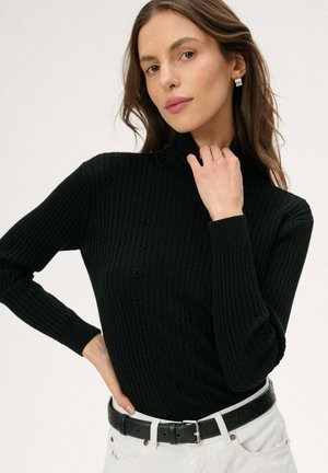 Kobieta ubrana w czarny prążkowany golf i białe spodnie z czarnym paskiem, pozująca z jedną ręką na biodrze, a drugą przy szyi.