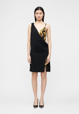 Jeune femme portant une robe portefeuille noire à imprimé baroque doré, talons hauts noirs, debout devant un fond gris clair uni.