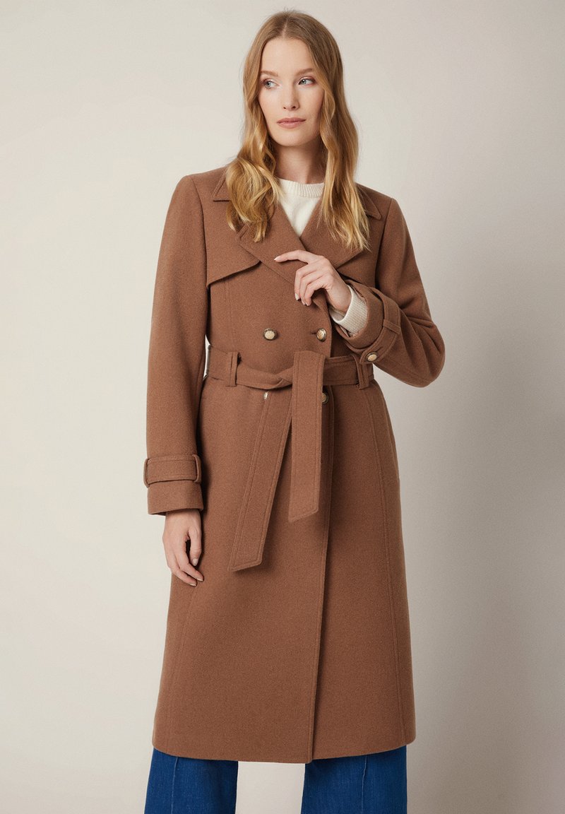 Brązowy płaszcz trench z mieszanki wełny, z podwójnym zapięciem na złote guziki, z paskiem w talii, szerokim kołnierzem i długimi rękawami, zestawiony z niebieskimi dżinsami.