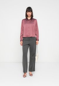 Blusa mauve a maniche corte con colletto e bottoni, abbinata a pantaloni grigi a gamba larga. Gli accessori includono una piccola borsa a tracolla bianca e tacchi.