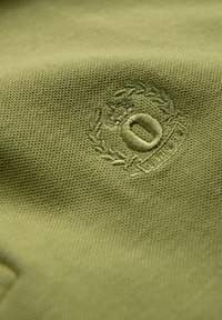 Gros plan sur un tissu vert texturé avec un emblème circulaire brodé présentant un "O" central entouré de feuilles de laurier et d'une bannière.