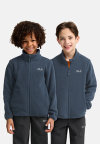 Due bambini indossano giacche di pile blu con colli alti e cerniere, dotate di un logo sul petto sinistro e abbinate a pantaloni neri.
