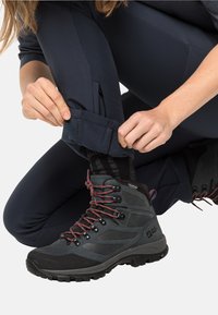 Bottes de randonnée noires avec des lacets rouges, des superpositions texturées et une semelle robuste. Une personne ajuste le poignet d'un pantalon sombre avec une fonctionnalité de zip.