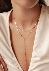 Gestapelte Halskette aus Roségold mit strukturierten, flachen Gliedern und einem Y-förmigen Anhänger, getragen mit einem cremefarbenen Oberteil mit subtiler Rippung.