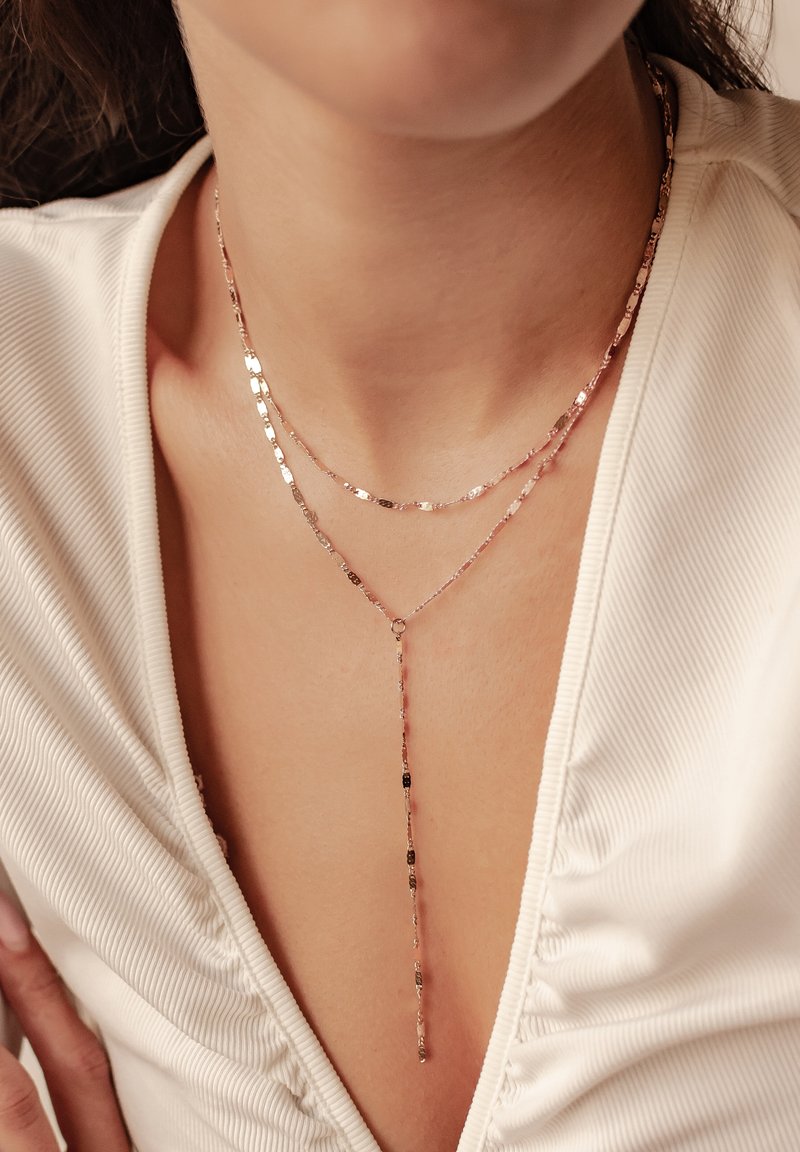 Gestapelte Halskette aus Roségold mit strukturierten, flachen Gliedern und einem Y-förmigen Anhänger, getragen mit einem cremefarbenen Oberteil mit subtiler Rippung.