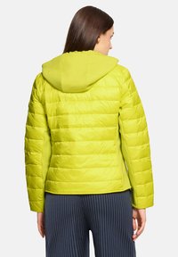 Limonkowo-zielona kurtka puchowa z kapturem, z pikowanym wzorem i ściągaczami na rękawach, zestawiona z granatowymi spodniami w paski.